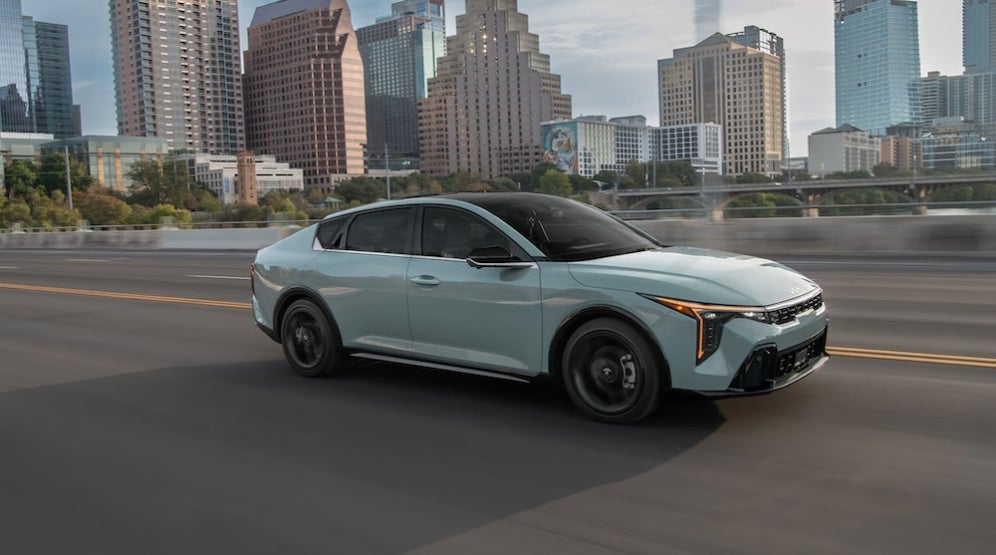 A light blue 2026 Kia K4 sedan driving down the highway with a cityscape in the background