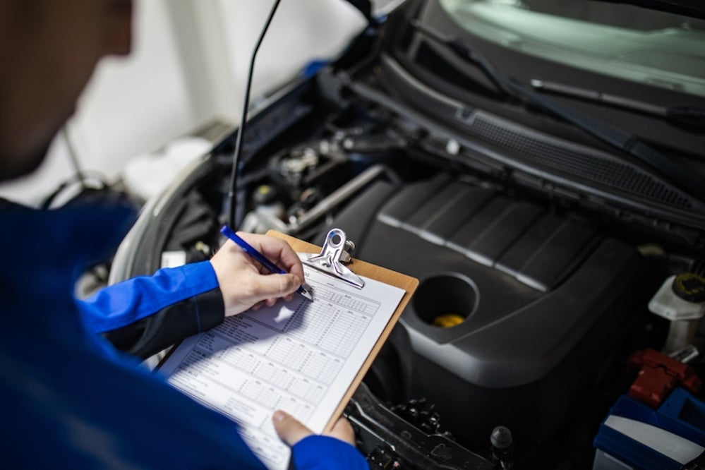 A mechanic in blue coveralls examining a vehicle’s engine and making notes on a clipboard