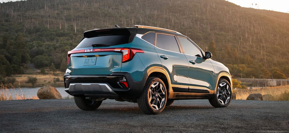 A turquoise 2026 Kia Seltos SUV parked in front of a body of water, with a hill in the background