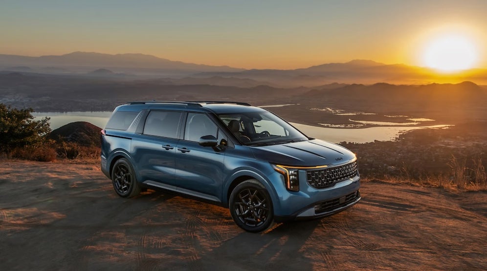 A blue 2027 Kia Carnival minivan parked on a dirt ledge at sunset