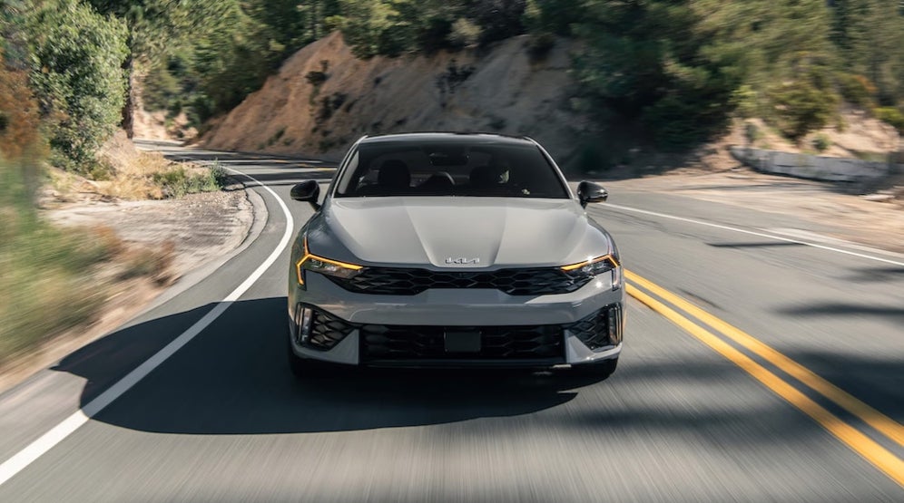 A gray 2026 Kia K5 sedan driving down a curving road